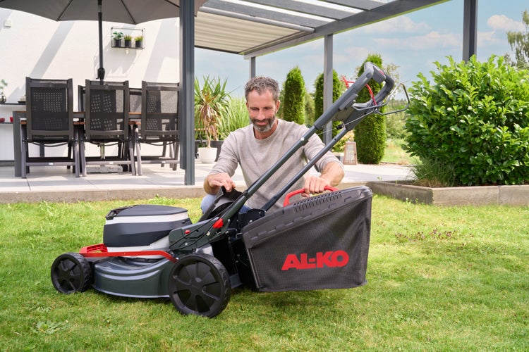 Ein Mann befestigt den Grasfangkorb an einem AL-KO Rasenmäher auf einem gepflegten Rasen. Im Hintergrund sind eine moderne Terrasse und Gartenmöbel.