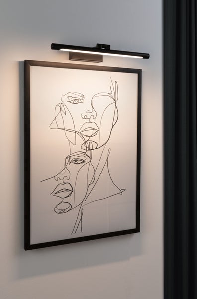 Schwarze LED-Bilderleuchte beleuchtet ein gerahmtes Kunstwerk mit Linienzeichnung an einer Wand.