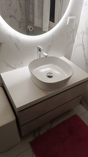 Modernes Badezimmer mit Waschtischunterschrank und Aufsatzwaschbecken vor einer Wand mit Marmorfliesen und beleuchtetem Rundspiegel.