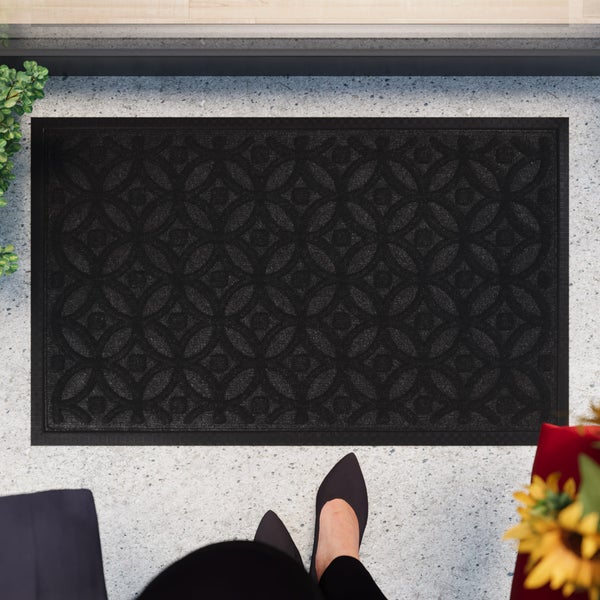 Dunkle Fußmatte mit geometrischem Kreismuster vor einem Hauseingang auf hellem Steinboden.