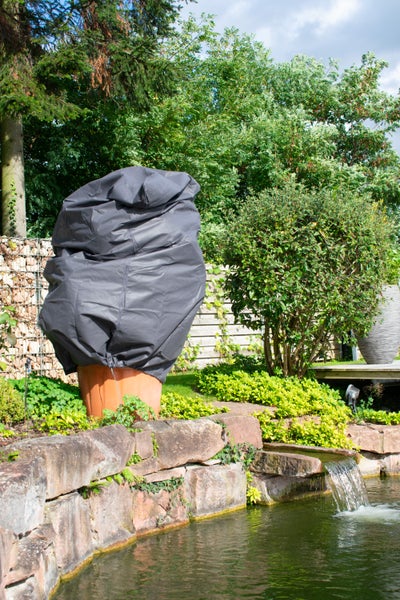 Eine Schutzhülle für Pflanzen steht in einem Garten neben einem Teich mit Steinen und grüner Bepflanzung.