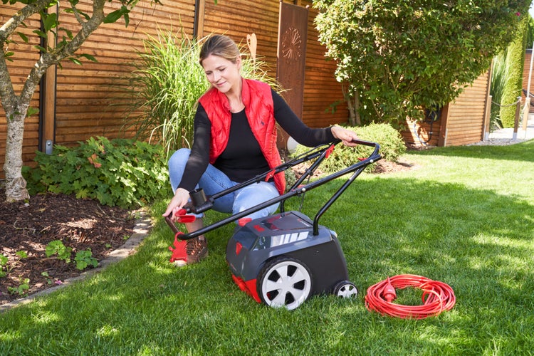 Eine Frau stellt den Holm eines Elektro-Vertikutierers auf einer Rasenfläche ein, daneben liegt ein rotes Verlängerungskabel.