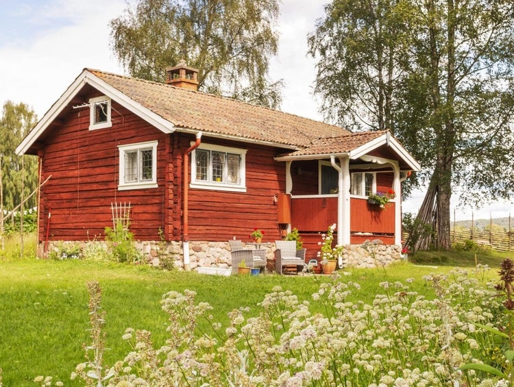 Rotes Holzhaus mit Veranda, weißen Fenstern und Ziegeldach in einem hellen Garten mit Wildblumen und grünem Rasen.