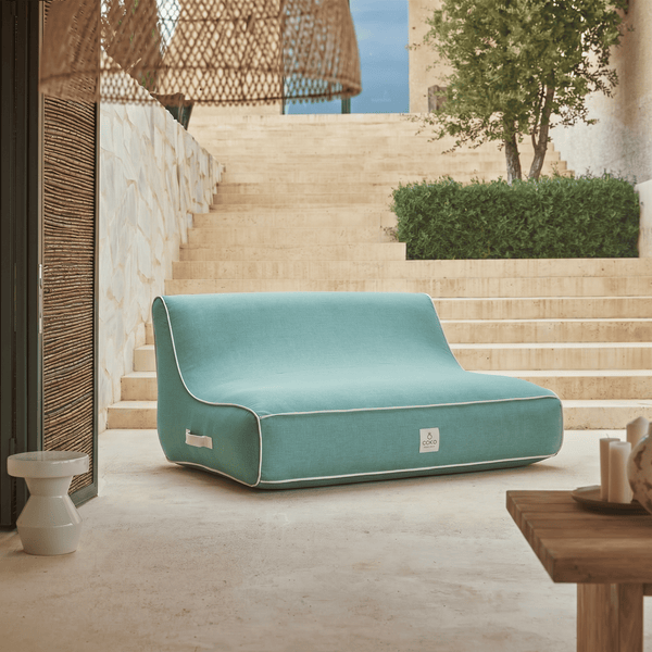 Modernes türkisblaues Outdoor-Sitzmöbel mit Coko Logo auf einer hellen, weitläufigen Steinterrasse mit Treppe und grüner Hecke.