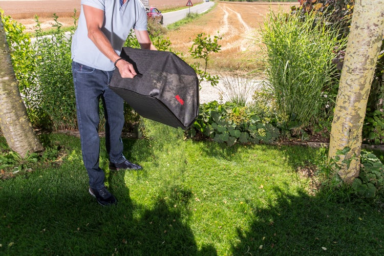 Person leert einen textilen Grasfangkorb für Rasenmäher der Marke Al-Ko über einer Rasenfläche in einem Garten aus.
