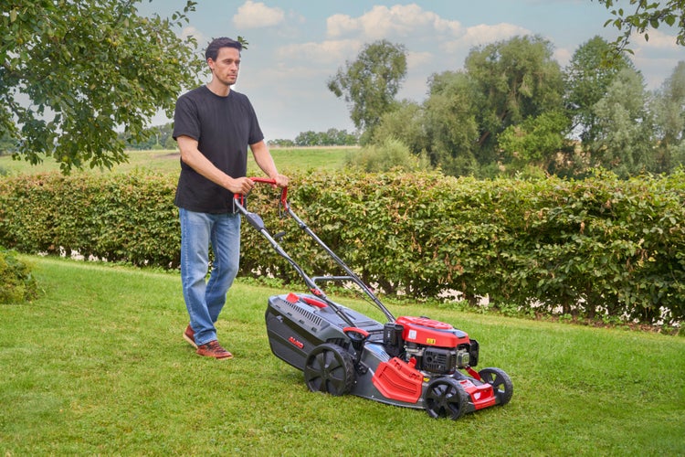 Ein Mann mäht Rasen mit einem AL-KO Benzinrasenmäher in einem Garten mit Hecke.
