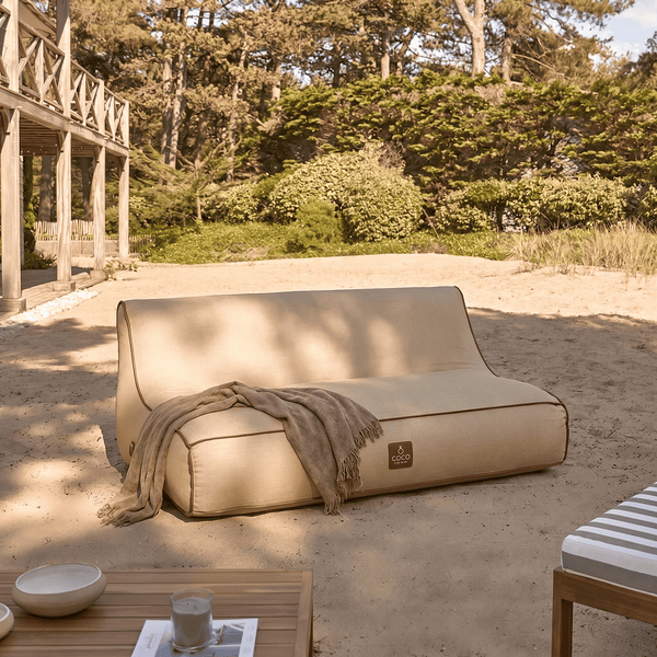 Modernes Outdoor-Lounge-Sofa mit dunkelbrauner Paspelierung und Decke auf Sand, im Hintergrund Bäume und eine Holzterrasse.
