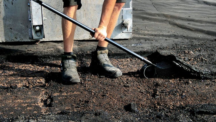 Person entfernt altes Bitumen oder Asphalt mit einem langstieligen Schaber auf einer Baustelle.