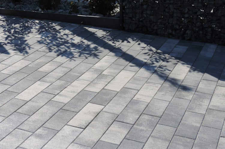 Pflastersteine aus Beton in Grau für Einfahrt oder Terrasse mit Baumschatten und Steinkorb im Hintergrund.