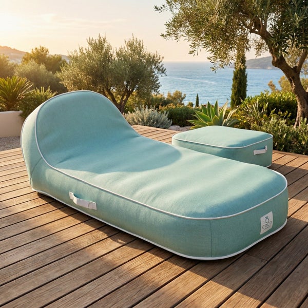 Modernes Set aus gepolsterter Sonnenliege und quadratischem Hocker mit Coco Lounge Logo auf einer Holzterrasse im Freien mit Küstenlandschaft.
