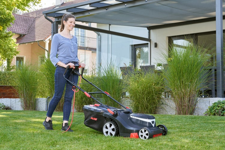 Eine Frau mäht mit einem elektrischen Rasenmäher der Marke AL-KO einen gepflegten Rasen im Garten vor einer modernen Terrasse.