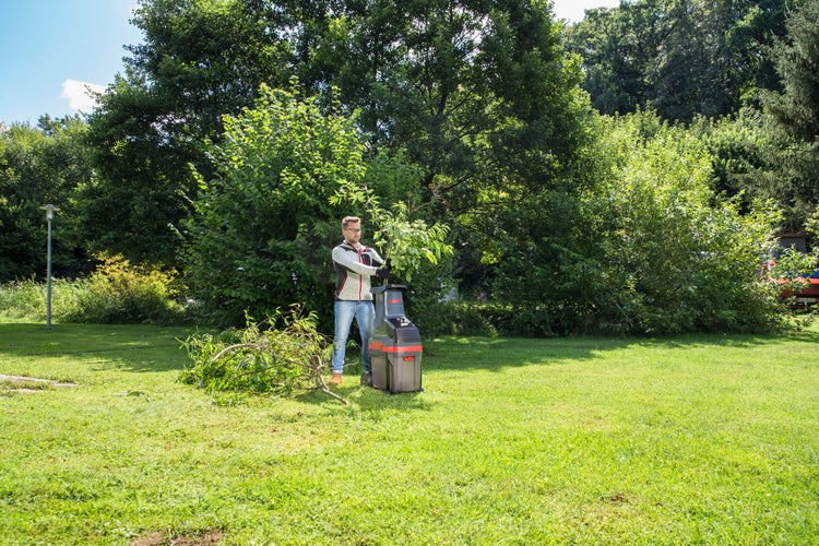 Ein Mann zerkleinert im Garten Äste mit einem elektrischen Messerhäcksler auf einer Rasenfläche vor Bäumen.