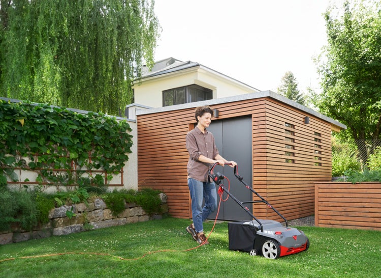 Eine Frau bedient einen Elektro-Vertikutierer auf einem grünen Rasen im Garten vor einem modernen Gartenhaus aus Holz.