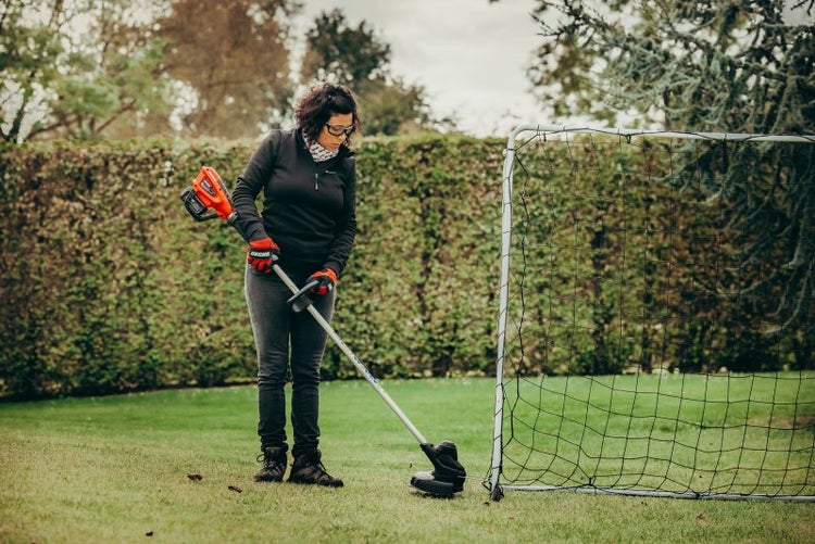 Frau arbeitet mit Akku-Rasentrimmer im Garten neben einem Fußballtor.