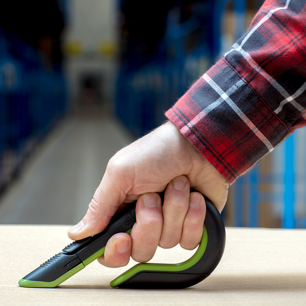 Hand in einem karierten Hemd benutzt ein ergonomisches Sicherheitsmesser zum Schneiden eines Kartons in einem Lager.