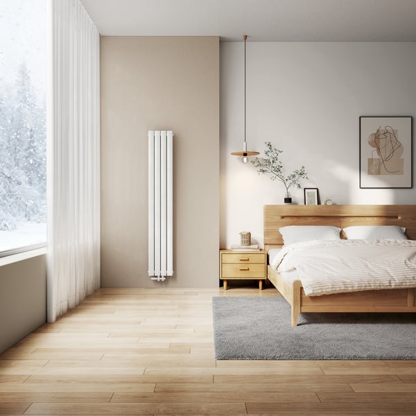 Ein moderner, vertikaler Röhrenheizkörper an einer Wand in einem hell eingerichteten Schlafzimmer mit Holzbett und Holzboden.