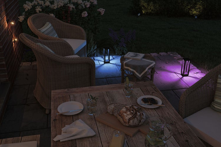 Abendliche Gartenszene mit Korbsesseln, Holztisch, Geschirr, Getränken und farbigen Gartenleuchten