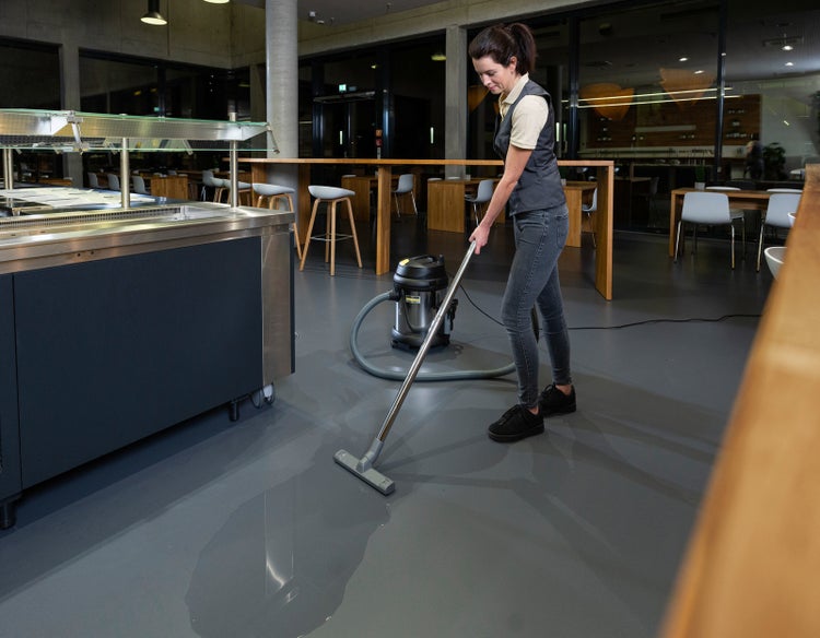 Eine Person reinigt mit einem Nass- und Trockensauger eine Wasserlache vom Boden in einer modernen Kantine.