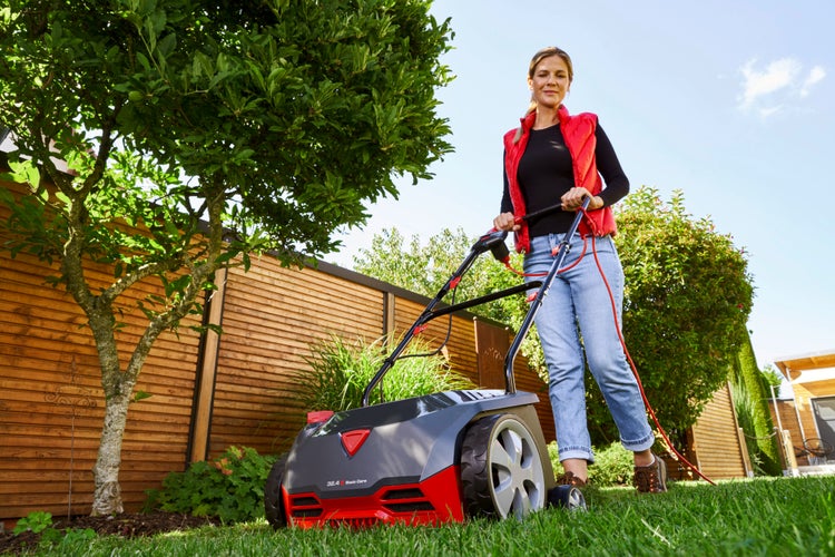 Eine Frau bedient einen Elektro-Vertikutierer auf einer grünen Rasenfläche im Garten.