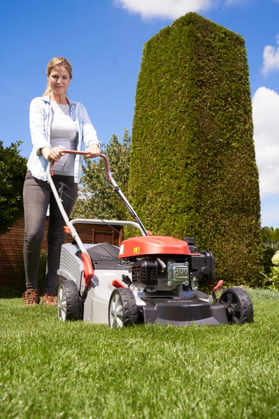 Eine Frau nutzt einen Benzinrasenmäher im Garten vor einer hohen Hecke.