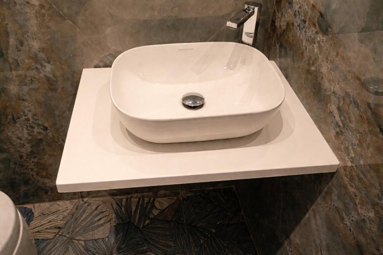 Weißes Aufsatzwaschbecken mit Armatur auf einer weißen Waschtischplatte vor einer Wand mit Natursteinoptik in einem modernen Badezimmer.