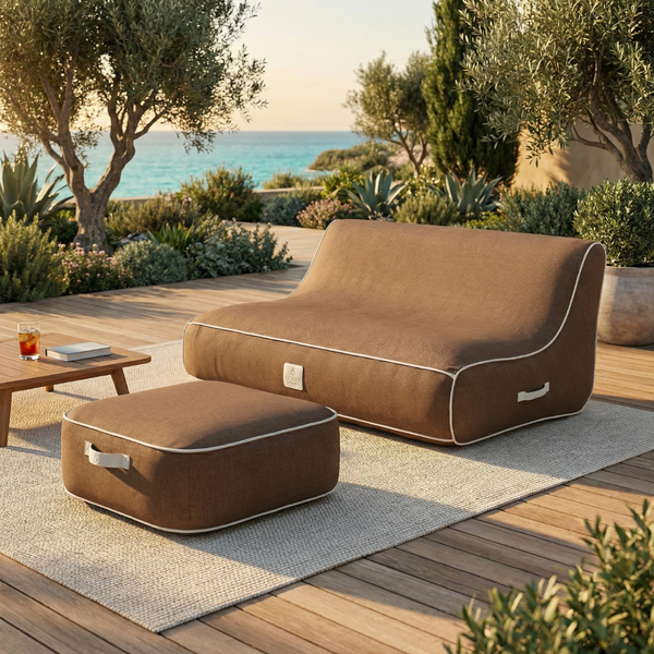 Modernes braunes Outdoor-Sitzsack-Sofa und passender Hocker auf einem hellen Teppich auf einer Holzterrasse mit Meerblick.
