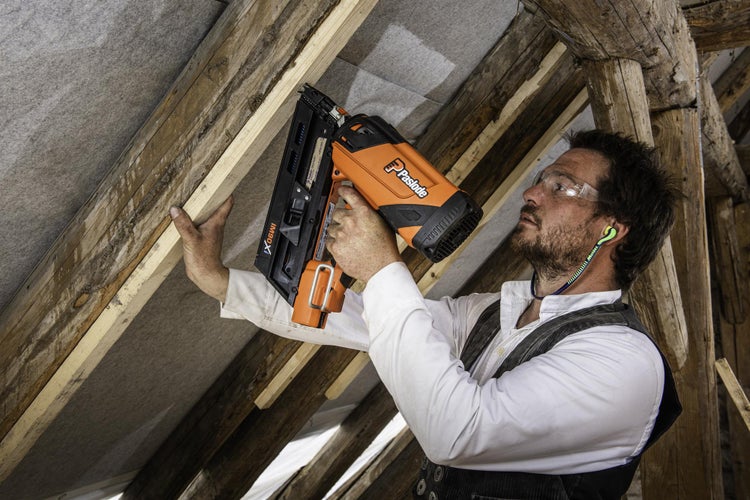 Ein Handwerker befestigt mit einem orangefarbenen Paslode Akku-Nagler eine Holzleiste an einem Dachstuhl.