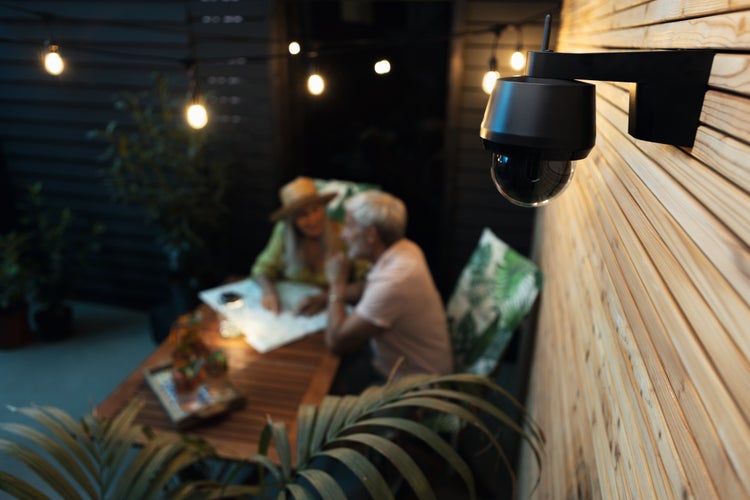 Schwarze Außenüberwachungskamera an einer Holzwand über einer beleuchteten Terrasse mit Personen bei Nacht.