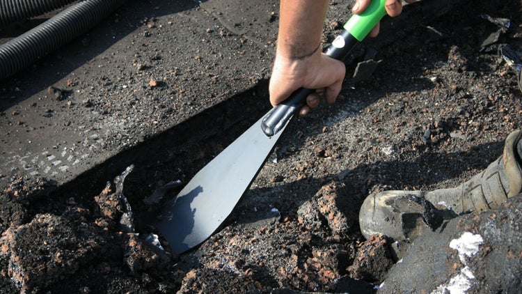 Ein Handwerker entfernt mit einem robusten Metallschaber alten Bitumenbelag von einer Baustellenoberfläche.