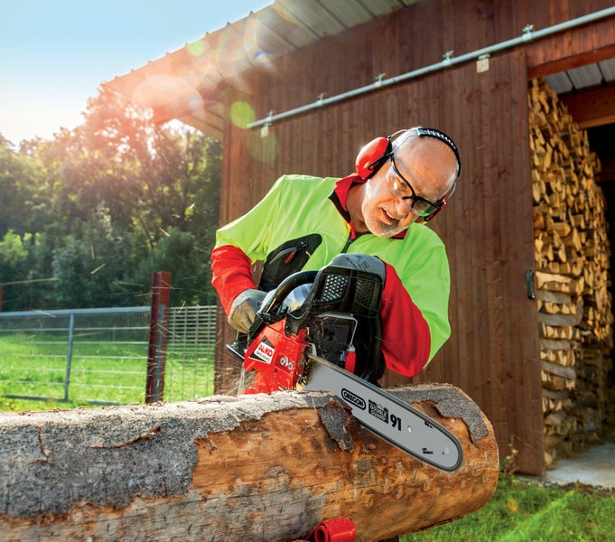 Ein Mann mit Schutzausrüstung schneidet einen Baumstamm mit einer AL-KO Kettensäge vor einem Holzschuppen.