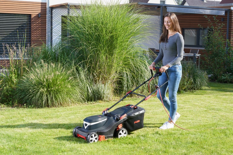 Eine Frau mäht mit einem Elektro Rasenmäher von AL-KO einen gepflegten Rasen vor einem modernen Wohnhaus.