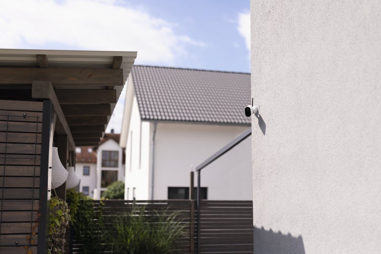 Weiße Außen-Überwachungskamera an einer Hauswand montiert, im Hintergrund ein Garten und ein Nachbarhaus.