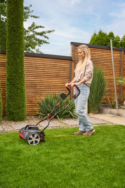 Eine Frau bedient einen Vertikutierer auf einer grünen Rasenfläche in einem Garten mit Holzzaun und Pflanzen.