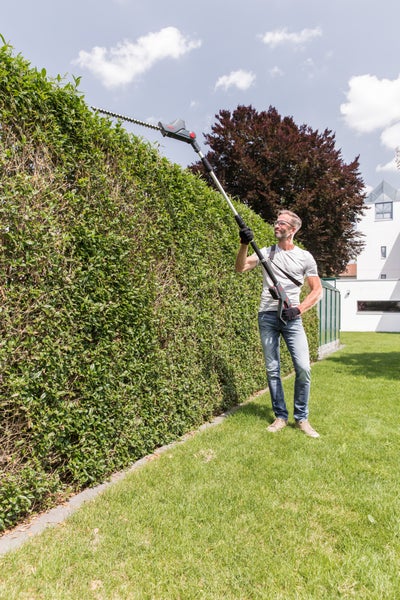Ein Mann schneidet eine hohe grüne Hecke im Garten mit einer Teleskop-Heckenschere.