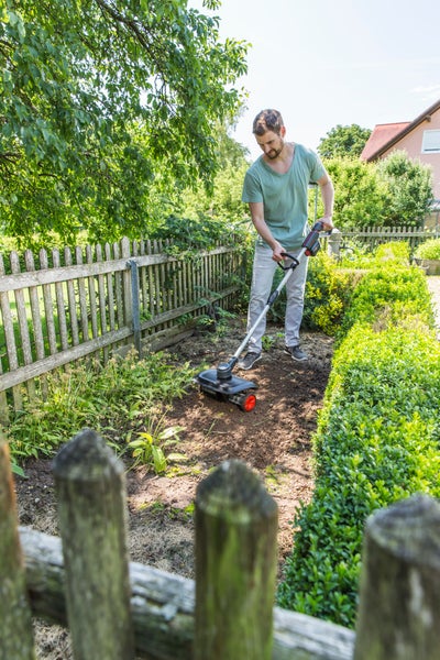 Ein Mann benutzt eine Akku-Bodenhacke zum Auflockern der Erde in einem Gartenbeet neben einem Holzzaun und einer Hecke.