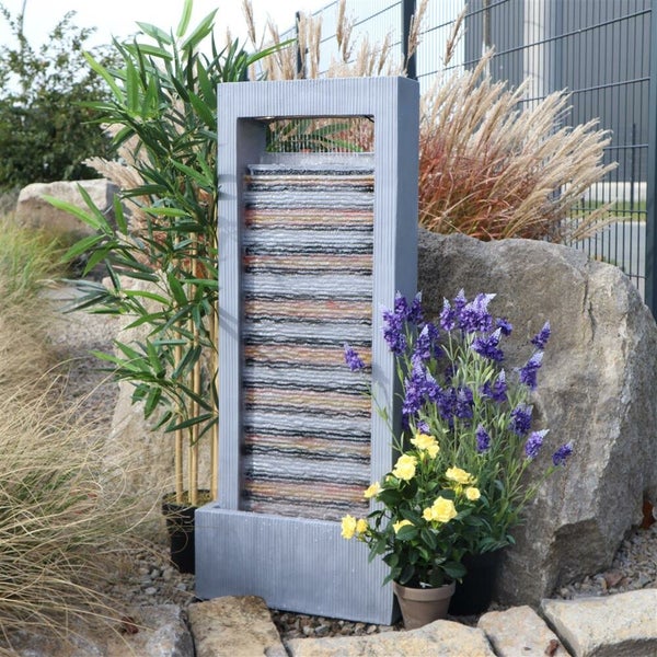 Wasserfallbrunnen mit rechteckigem Rahmen, umgeben von Pflanzen und Steinen im Garten.