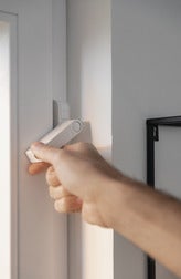 Eine Hand betätigt einen weißen Fensterkontakt an einem Fensterrahmen.