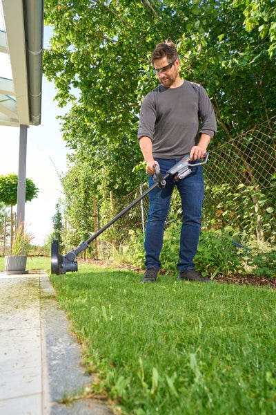 Ein Mann benutzt einen Akku-Rasentrimmer zum Schneiden der Rasenkante neben einem gepflasterten Weg im Garten.