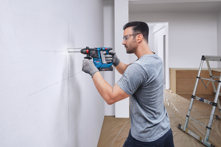Mann bohrt mit einem Bosch Professional Akku-Bohrhammer in eine weiße Wand bei Renovierungsarbeiten, trägt Schutzbrille und Handschuhe.