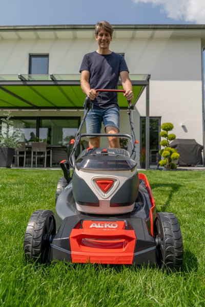 Ein Mann mäht den Rasen mit einem akkubetriebenen AL-KO Rasenmäher Comfort 46.2 Li SP vor einem modernen Wohnhaus mit Terrasse.