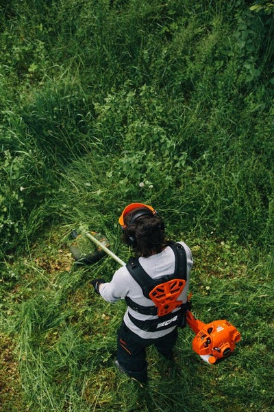 Eine Person schneidet mit einem Rasentrimmer hohes Gras. Die Person trägt Schutzkleidung.