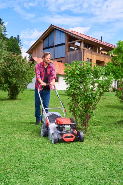 Ein Mann mäht Rasen mit einem Benzinrasenmäher in einem Garten vor einem Holzhaus mit Giebeldach bei Sonnenschein.