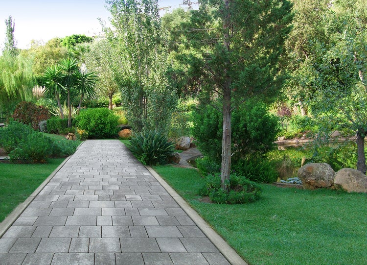 Moderner Gartenweg aus rechteckigen Betonpflastersteinen in einer gepflegten Gartenanlage mit Bäumen, Sträuchern, Rasen und einem Teich.