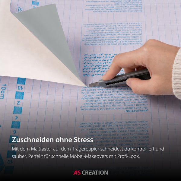 Eine Hand schneidet eine Klebefolie mit einem Cuttermesser entlang des Maßrasters auf dem Trägerpapier zu. AS Creation Logo.