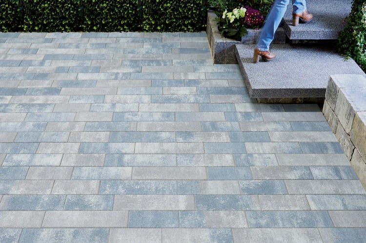 Terrasse mit grau melierten Pflastersteinen im rechteckigen Format, angrenzenden Blockstufen und einer grünen Hecke im Hintergrund.