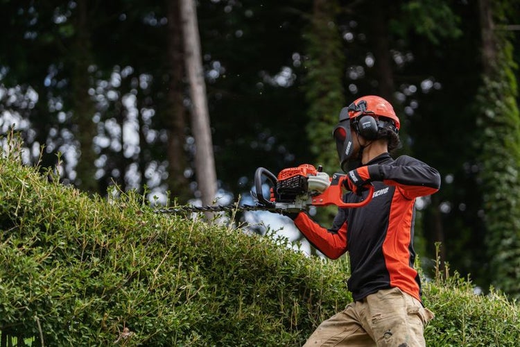 Eine Person, die eine Heckenschere mit Schutzausrüstung benutzt, um eine Hecke zu schneiden.