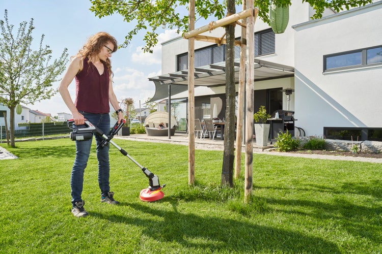 Eine Frau nutzt einen Akku Rasentrimmer für die Gartenarbeit im Garten eines modernen Einfamilienhauses.