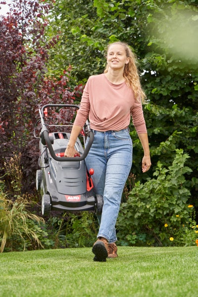 Eine Frau trägt einen Elektro-Rasenmäher von Alko Logo über einen Rasen im Garten vor einer Hecke.