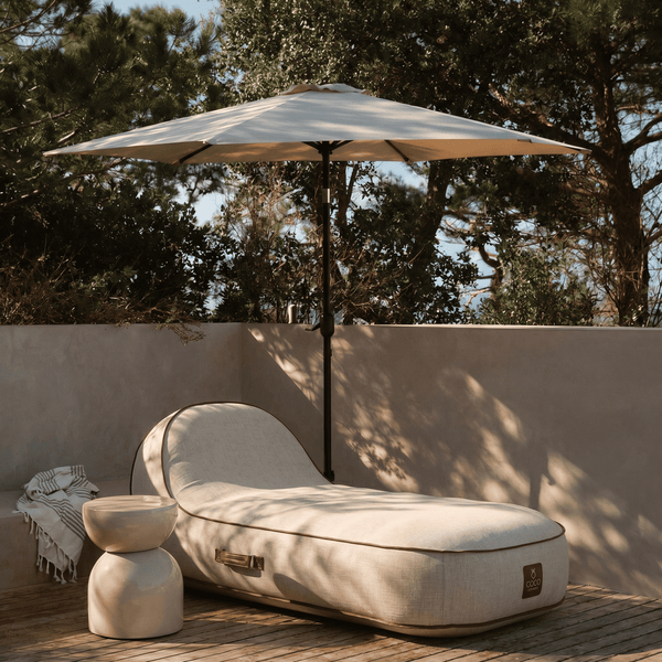 Beige Sitzsack-Gartenliege mit Coco Lounge Logo auf Steinterrasse am Pool in hellem Garten mit Holzzaun, Sonnenschirm und Pflanzen.