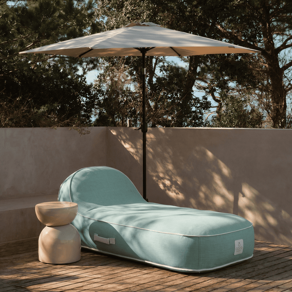 Gepolsterte Sonnenliege im Sitzsack-Stil mit Beistelltisch und Sonnenschirm auf einer Holzterrasse im Garten.